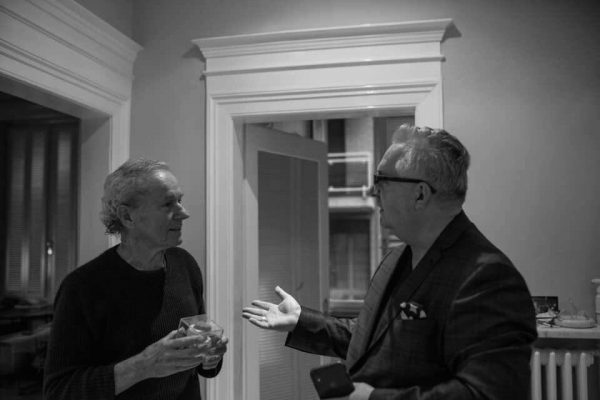 Two men engaged in conversation, one holding a drink, in a well-lit interior with doorways and a modern aesthetic, reflecting a social or professional setting.