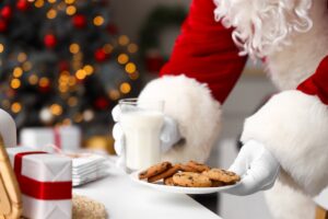 Santa Claus in a red suit holding a glass of milk and a plate of cookies, with a festive background of a Christmas tree adorned with lights and ornaments.