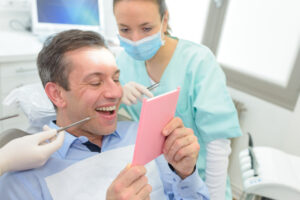 Man smiling while looking at a mirror in a dental office, with dental professionals assisting, showcasing the positive experience of cosmetic dentures.