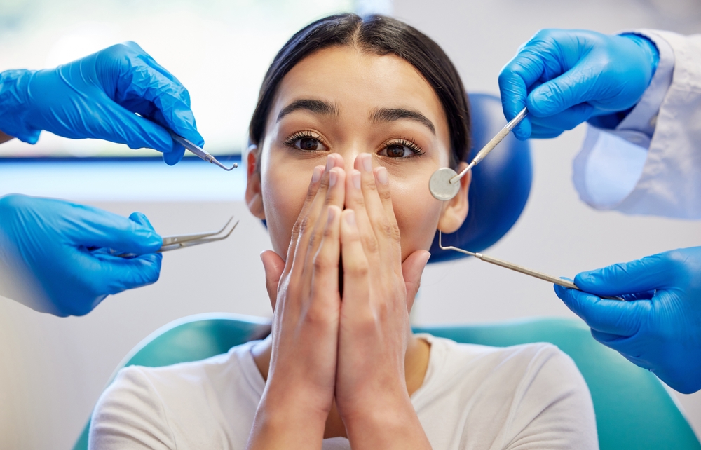 Woman in dental chair covering her face with hands, showing signs of anxiety, surrounded by dental tools and gloved hands of dental professionals, illustrating dental anxiety before an appointment.