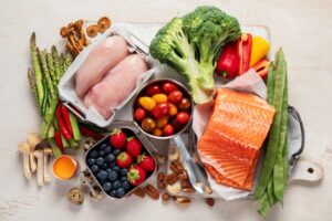 Nutrient-dense foods for recovery including salmon, chicken, broccoli, bell peppers, tomatoes, and berries, arranged on a wooden surface, emphasizing healthy eating during oral health recovery.